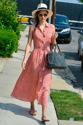 A woman in a red patterned dress, sun hat, sunglasses, and sandals walks on a sidewalk carrying a black bag.