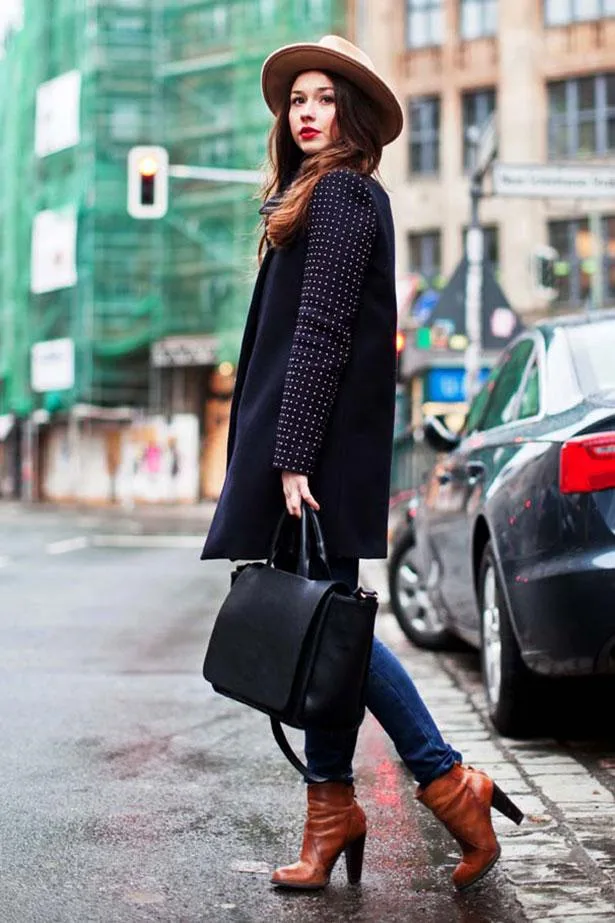 Woman in a stylish coat and hat, holding a black bag, stands on a city street corner near parked cars.