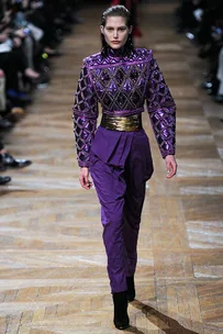 Model walks runway in a purple, embellished jumpsuit with gold belt, surrounded by audience on wooden floor.