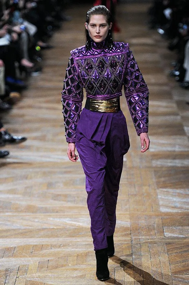 Model walks runway in a purple, embellished jumpsuit with gold belt, surrounded by audience on wooden floor.