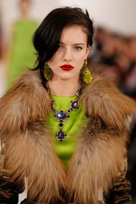 Model wearing fur coat with statement earrings and necklace, set against a blurred runway backdrop.