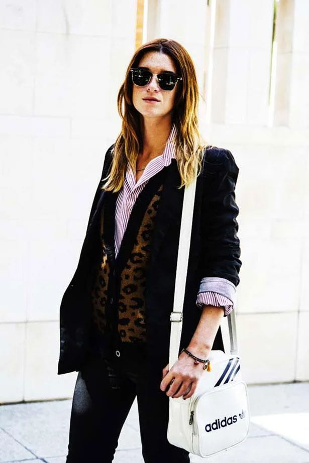 Person wearing sunglasses, striped shirt, leopard print vest, black blazer, carrying white Adidas bag.