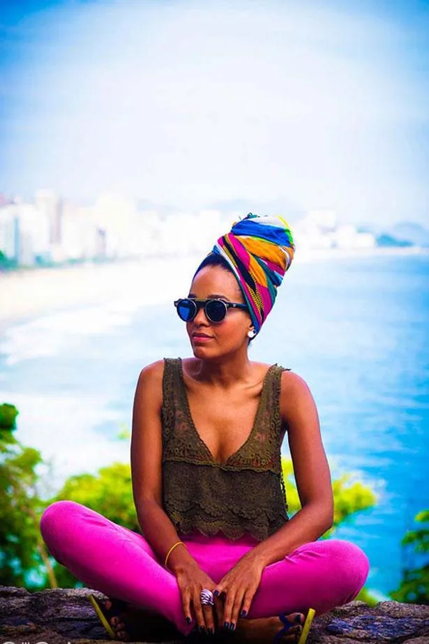 Woman in colorful headwrap and sunglasses sitting cross-legged near a coastal view.