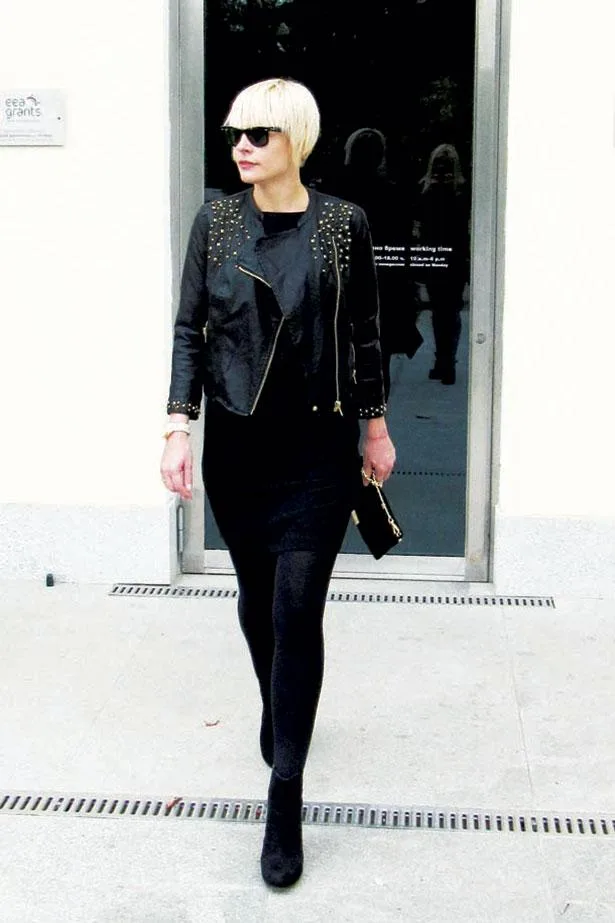 Woman in black studded jacket and sunglasses walking, holding a black clutch, against a glass door background.