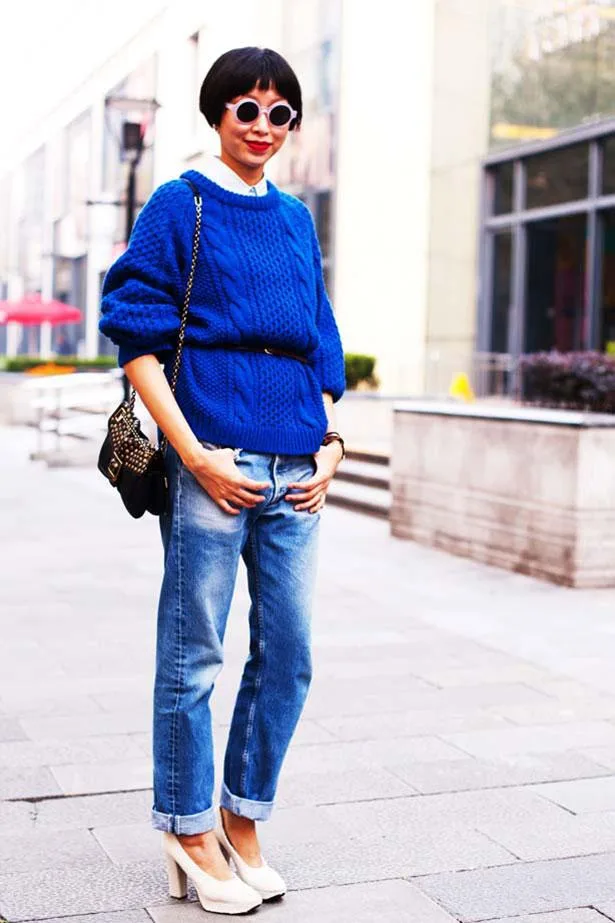 Woman in blue sweater, cuffed jeans, and heels stands on street with a bag, wearing round sunglasses and short haircut.