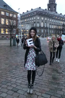 Woman in floral dress and beanie holding letter 'E' on cobblestone street in front of historic building.