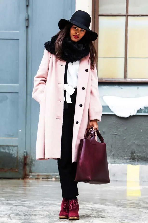 Woman in pink coat, black hat, and scarf holding burgundy bag, wearing red boots, standing on wet street.