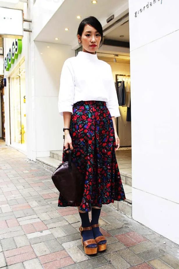 A woman in a white top and floral skirt stands on a Hong Kong street, holding a dark bag, wearing platform sandals.