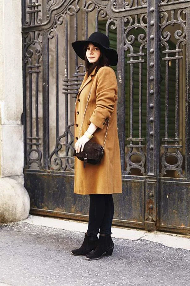 Woman in a brown coat and black hat standing in front of ornate metal gates.