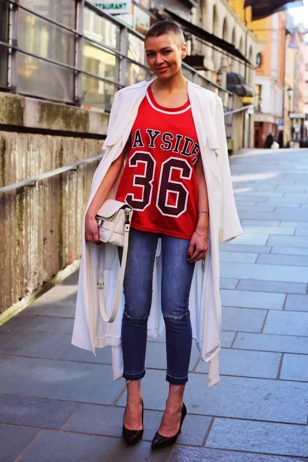 Woman in street style outfit: red jersey, white coat, jeans, and black heels, posing on a cobblestone street.