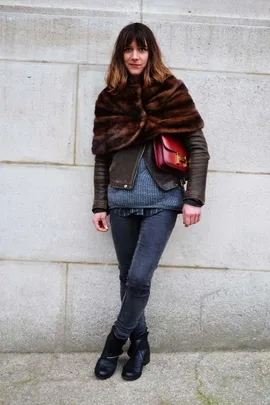 Street style: person in a fur wrap, leather and gray layered outfits with a red bag, standing against a light wall.