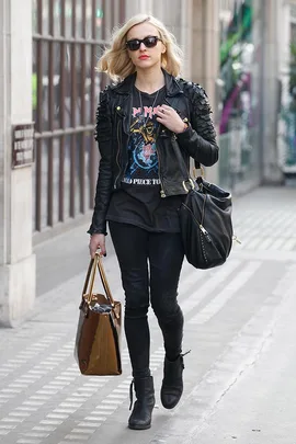 A woman in a black leather jacket and sunglasses walks on a sidewalk carrying two bags.