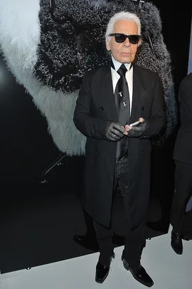 A man in a black suit, sunglasses, and gloves stands in front of a large black-and-white textured artwork.