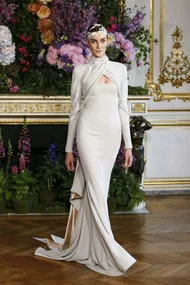 Model in an elegant white fantasy wedding dress with floral headpiece, standing in a lavish room with floral decor.