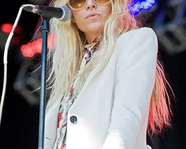 A woman in sunglasses and white suit sings into a microphone on stage with colorful lights in the background.