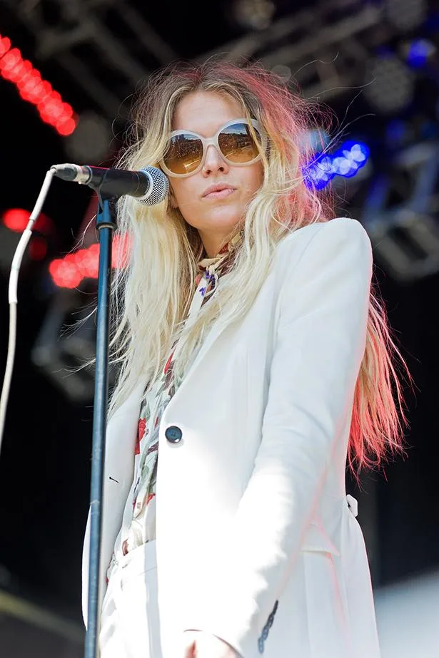 A woman in sunglasses and white suit sings into a microphone on stage with colorful lights in the background.