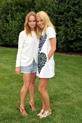 Gwyneth Paltrow and Stella McCartney posing together outdoors on grass with green foliage in the background.