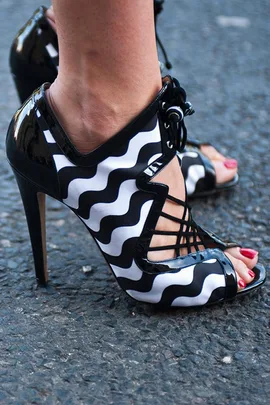 Black and white high-heeled shoes with wavy pattern, worn on a textured outdoor surface.