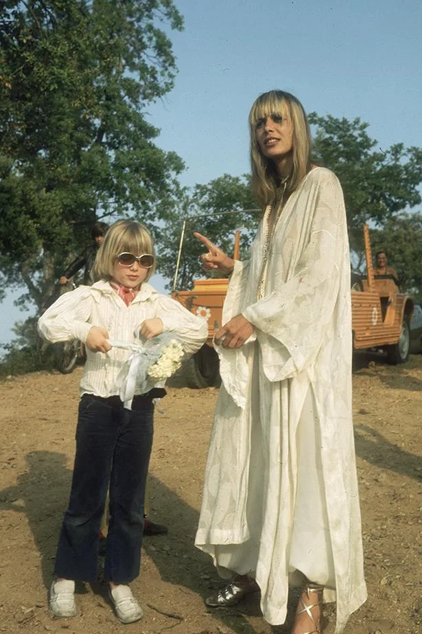 Woman in white dress with blond child holding flowers outdoors, near a vintage orange car under trees.