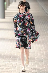 Male model in floral-patterned black outfit and sandals walks on a runway holding a matching hat.