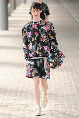 Male model in floral-patterned black outfit and sandals walks on a runway holding a matching hat.