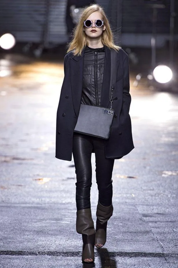 Model wearing black leather jacket, navy coat, sunglasses, crossbody bag, and gray peep-toe boots on a wet runway.