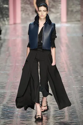 Model walking runway in a black outfit with long, flowing pants and a dark blue vest.