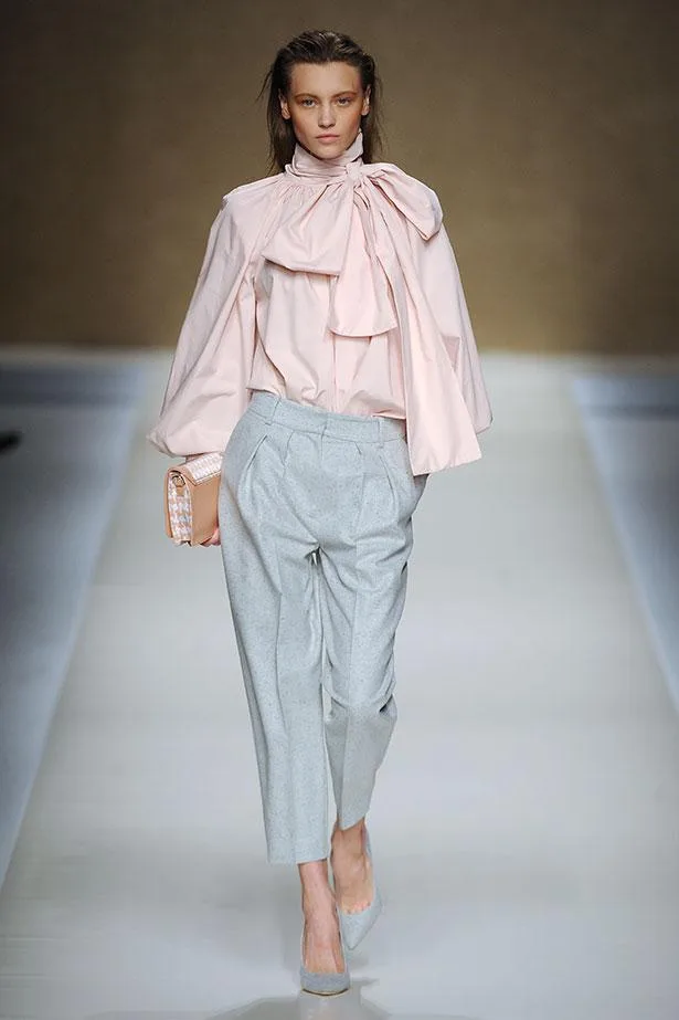 Model wearing a pink blouse with a large bow and gray trousers on the runway, holding a clutch.