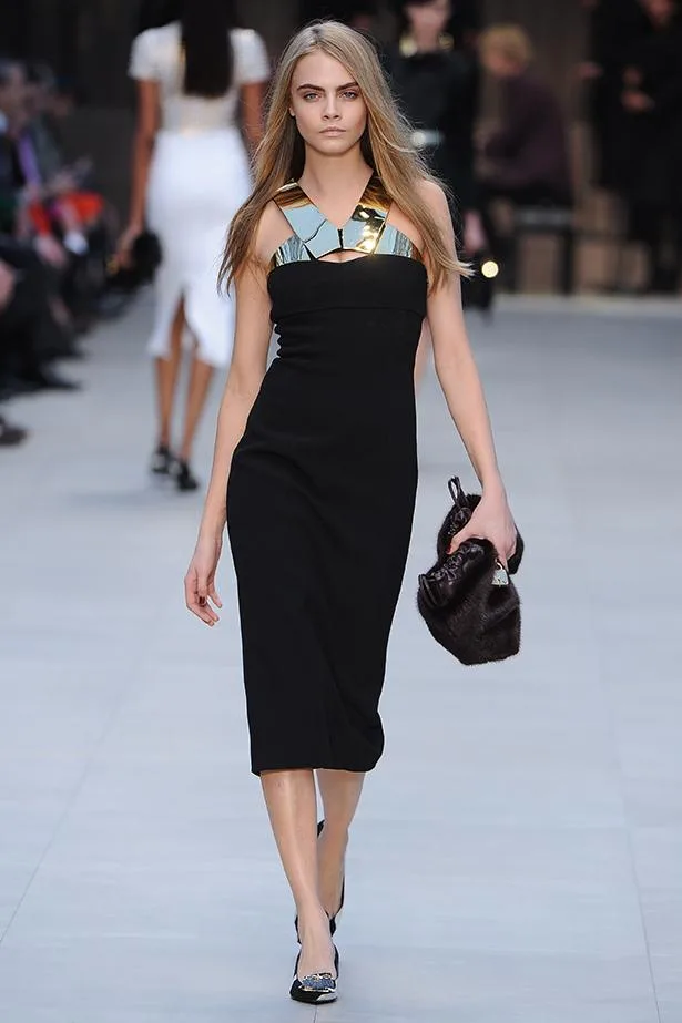 Model walking runway in a black dress with metallic neckline, holding a handbag, in a fashion show setting.