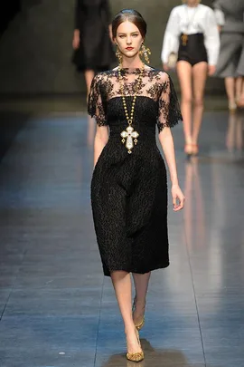 Model walks runway in a black lace dress with gold cross necklace, high bun, and gold shoes at fashion show.