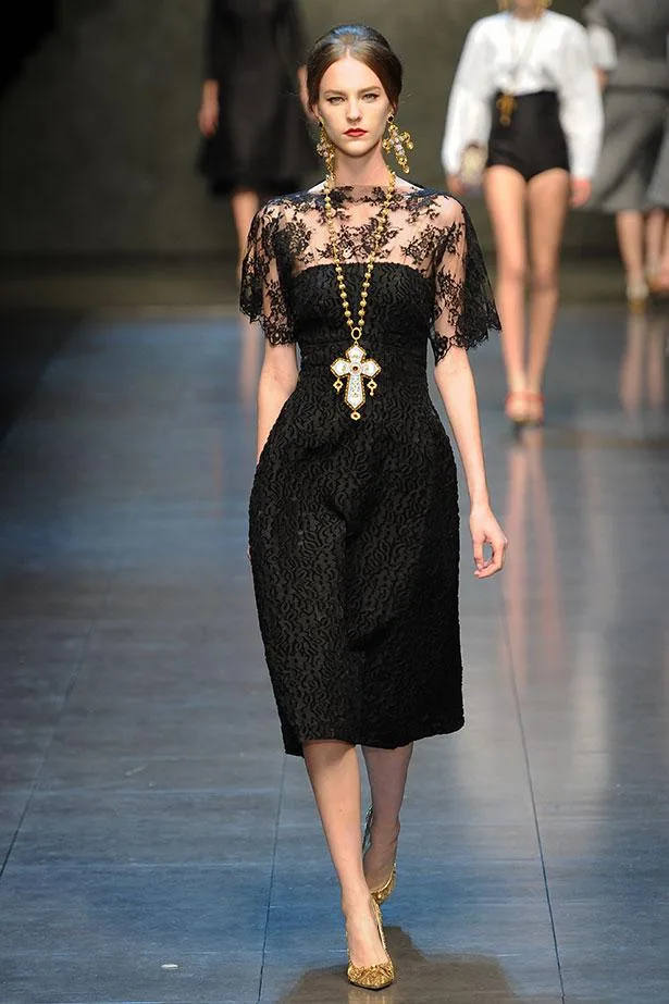 Model walks runway in a black lace dress with gold cross necklace, high bun, and gold shoes at fashion show.