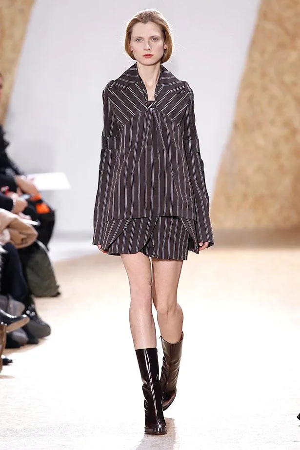 Model in striped brown outfit and knee-high boots walking on a runway during a fashion show.