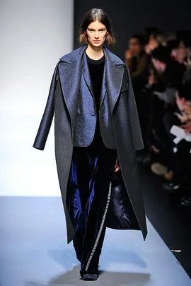 Model walks runway wearing oversized coat and velvet pants during a fashion show.