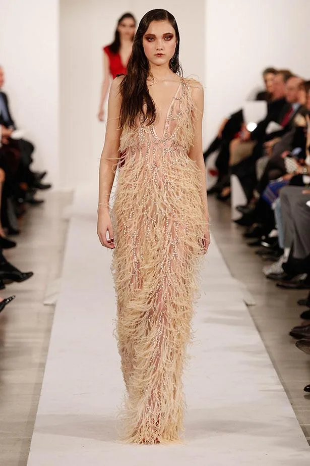 Model walking runway in a beaded, feathered gown with a deep neckline, against an audience background.