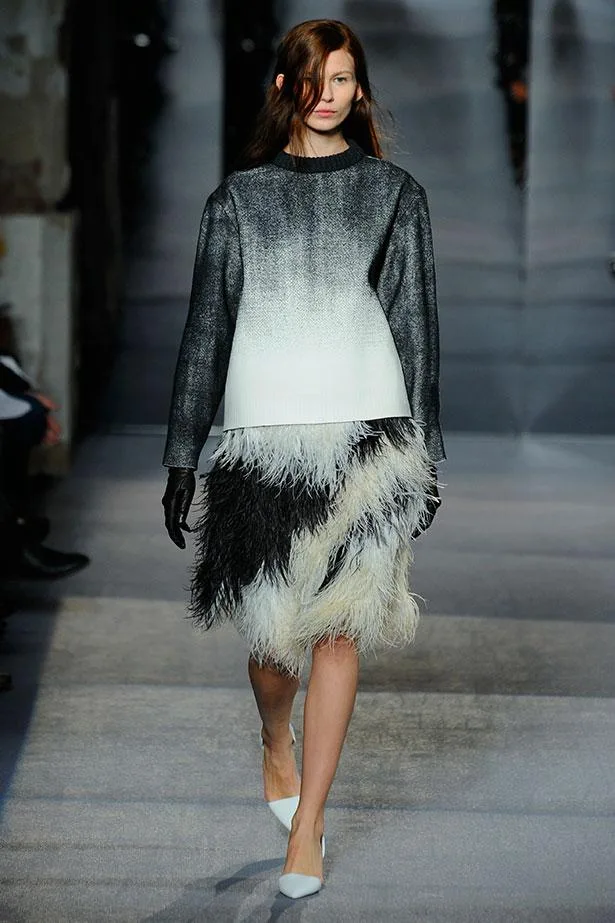 Model walking runway in ombre gray top with a black-and-white feathered skirt, wearing black gloves and white heels.