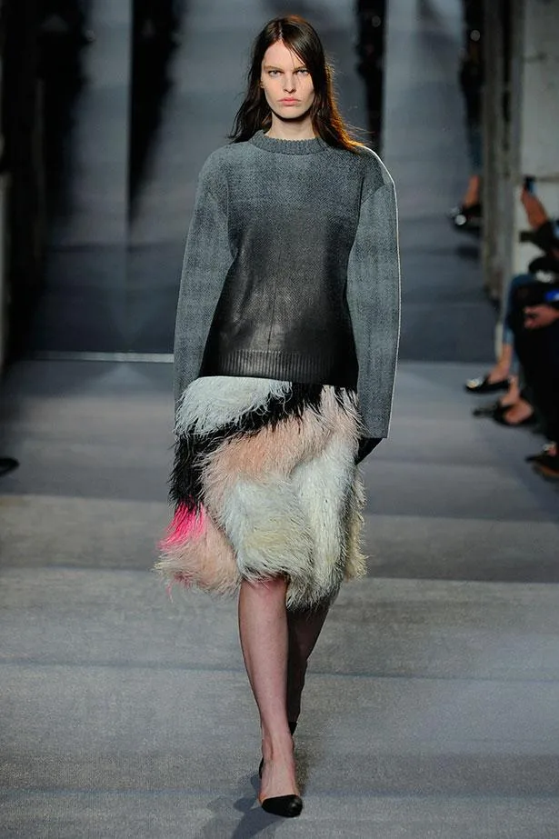 Model in a gray sweater and colorful feather skirt walking on a runway.