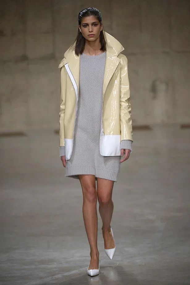 Model wearing a gray knit dress and a shiny beige coat, walking on a runway.