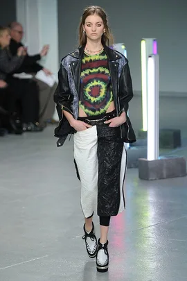 Model on runway in punk-inspired outfit: tie-dye shirt, leather jacket, black and white skirt, and creeper shoes.