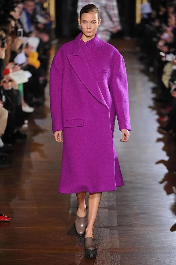 A model wearing a bold purple coat walks down the runway at a fashion show, surrounded by an audience.