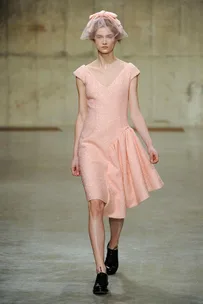 Model in a pastel pink dress with a matching headpiece walks on a concrete runway.