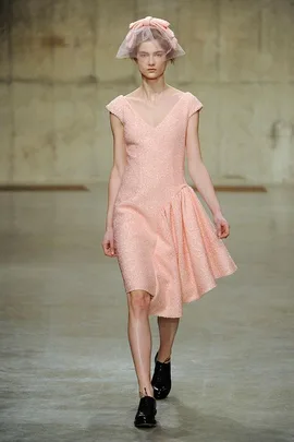 Model in a pastel pink dress with a matching headpiece walks on a concrete runway.