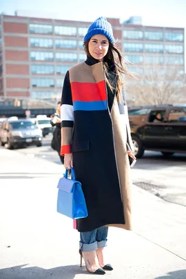 Woman in colorful coat, blue beanie, holding a blue handbag on a city street.