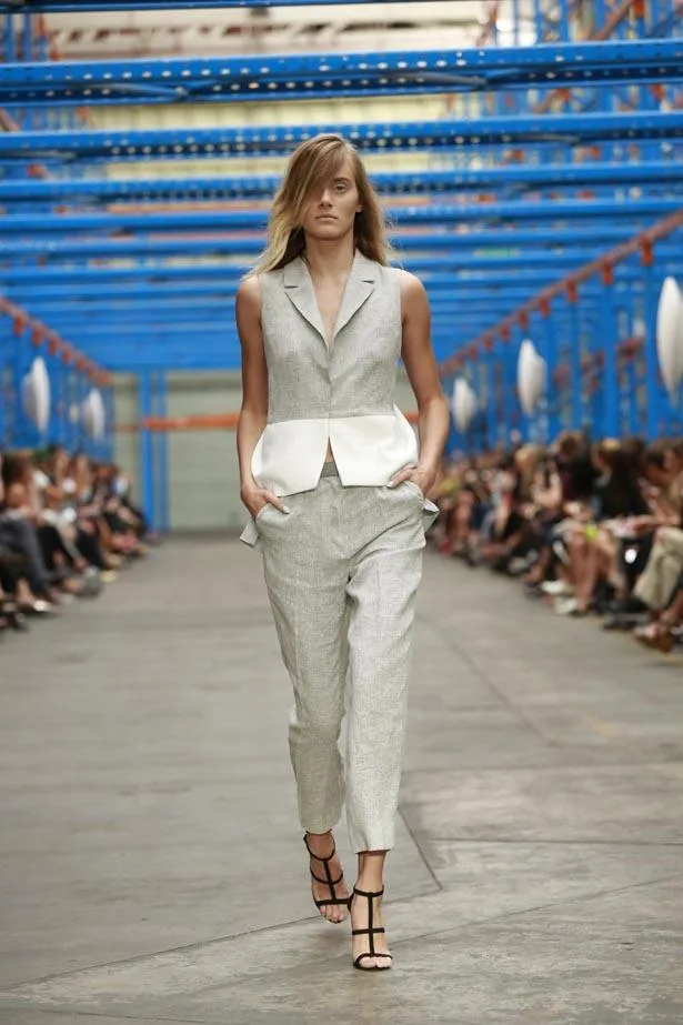 Model walking on a runway in a sleeveless gray vest and pants with black sandals, surrounded by an industrial setting.