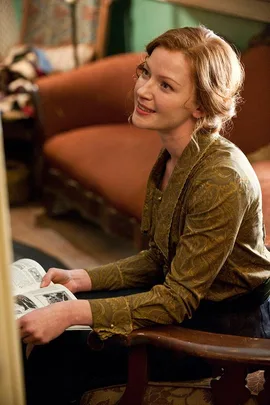 A woman in period attire sits in a vintage chair, holding an open book, smiling, in an early 20th-century setting.