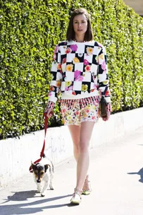 Woman in floral outfit walks a small dog on leash, against a green hedge background.