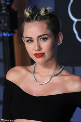 A person with short blonde hair in double buns, wearing a black off-shoulder top and a diamond necklace.