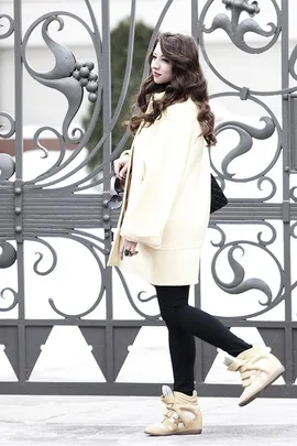 Woman in a cream coat and beige sneakers poses in front of an ornate metal gate on a snowy day.