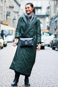 Woman in long green coat with crossbody bag, smiling on a cobblestone street; urban fashion street style.