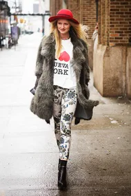 Woman in a fur coat, red hat, "I ♥ Nueva York" shirt, floral pants, walking on a city sidewalk.