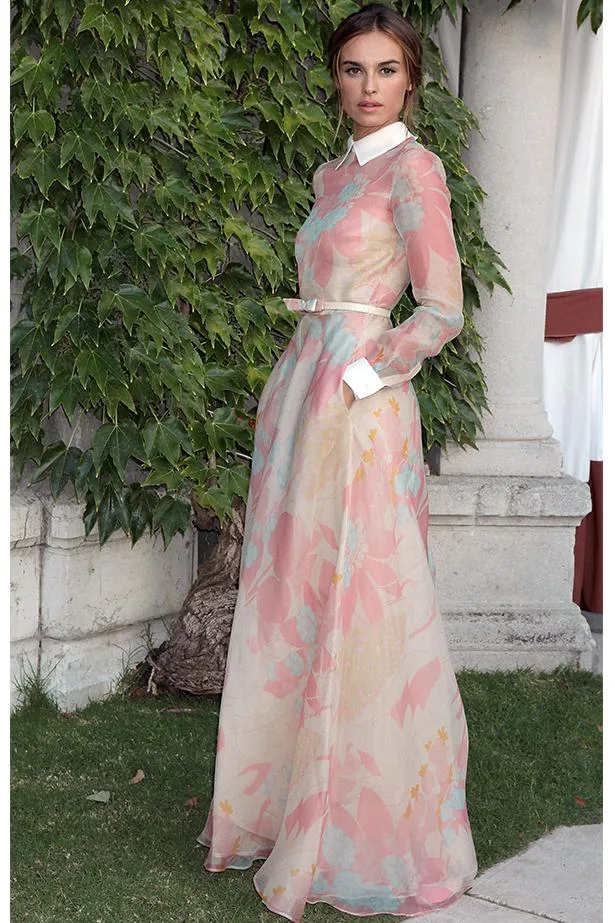 Woman in a sheer pastel floral gown with long sleeves and a white collar, standing in front of ivy-covered wall.